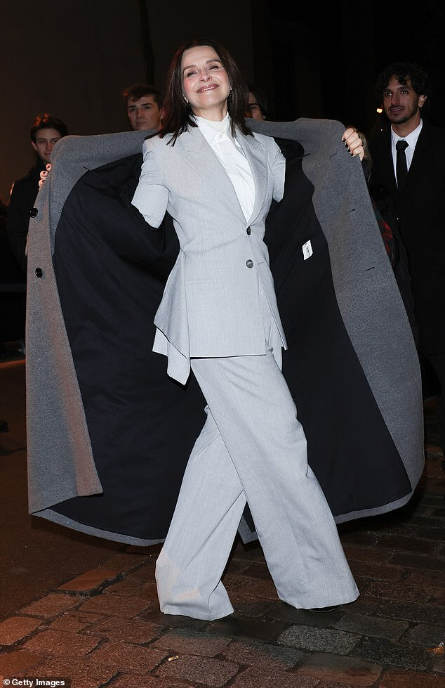 Жюльет Бинош расстегивает пальто и демонстрирует шикарный серый брючный костюм по прибытии на показ AMI Alexandre Mattiussi на Неделе моды в Париже.